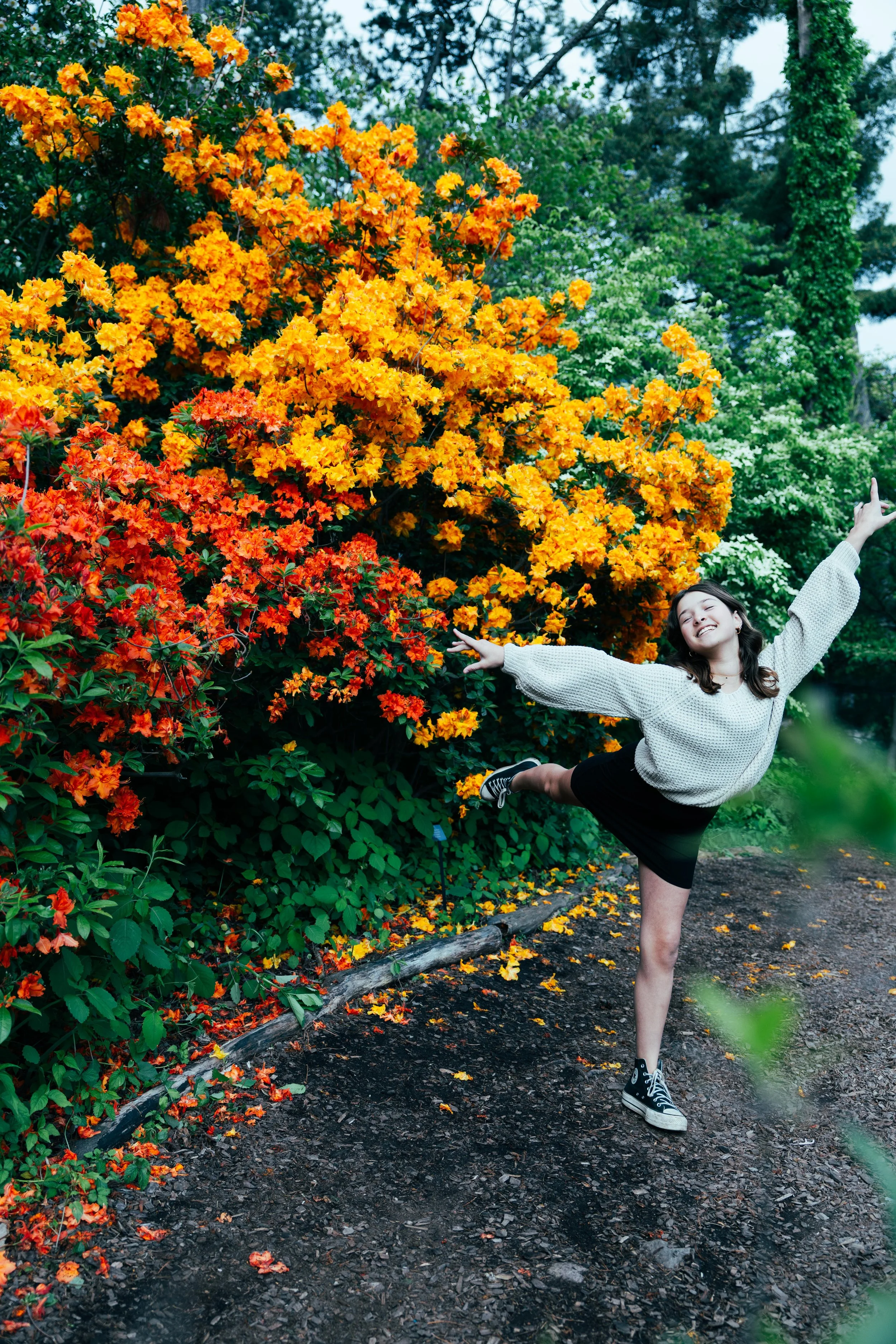 A woman with dark hair, wearing a gray sweater, black shorts, and black sneakers, is joyfully jumping with arms wide open in front of a large bush of bright orange and red flowers and green trees in the background, on a dirt path.