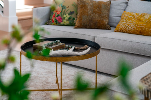 Living room sofa with colorful pillows and a round black and gold coffee table with decorative items, in front of a window.