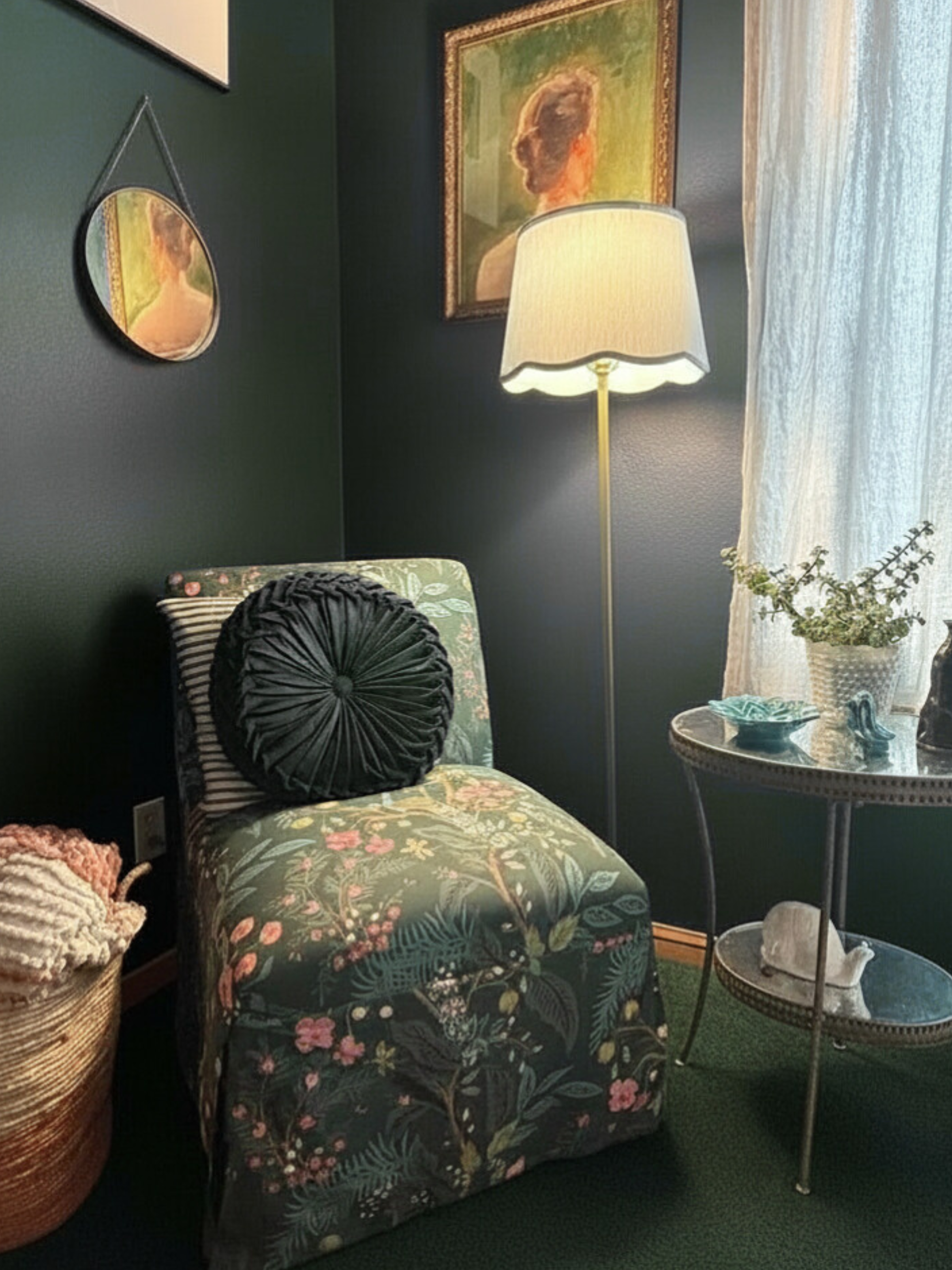 A cozy corner with a floral upholstered chair, a round black pillow, a tall floor lamp, a small side table with decorative items and a potted plant, framed artwork, and a mirror on a dark green wall near a window with sheer curtains.