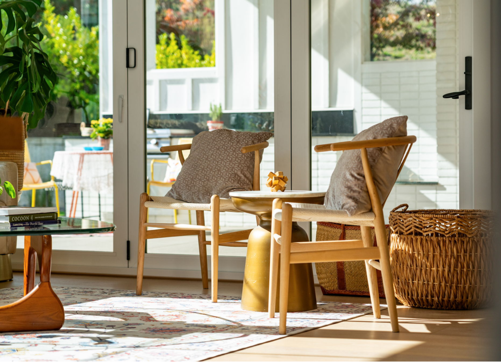 Sunlit living room with two wooden chairs with cushions, a gold side table, a basket, books, and a patterned rug, with a large glass door leading to an outdoor patio with yellow chairs and greenery.