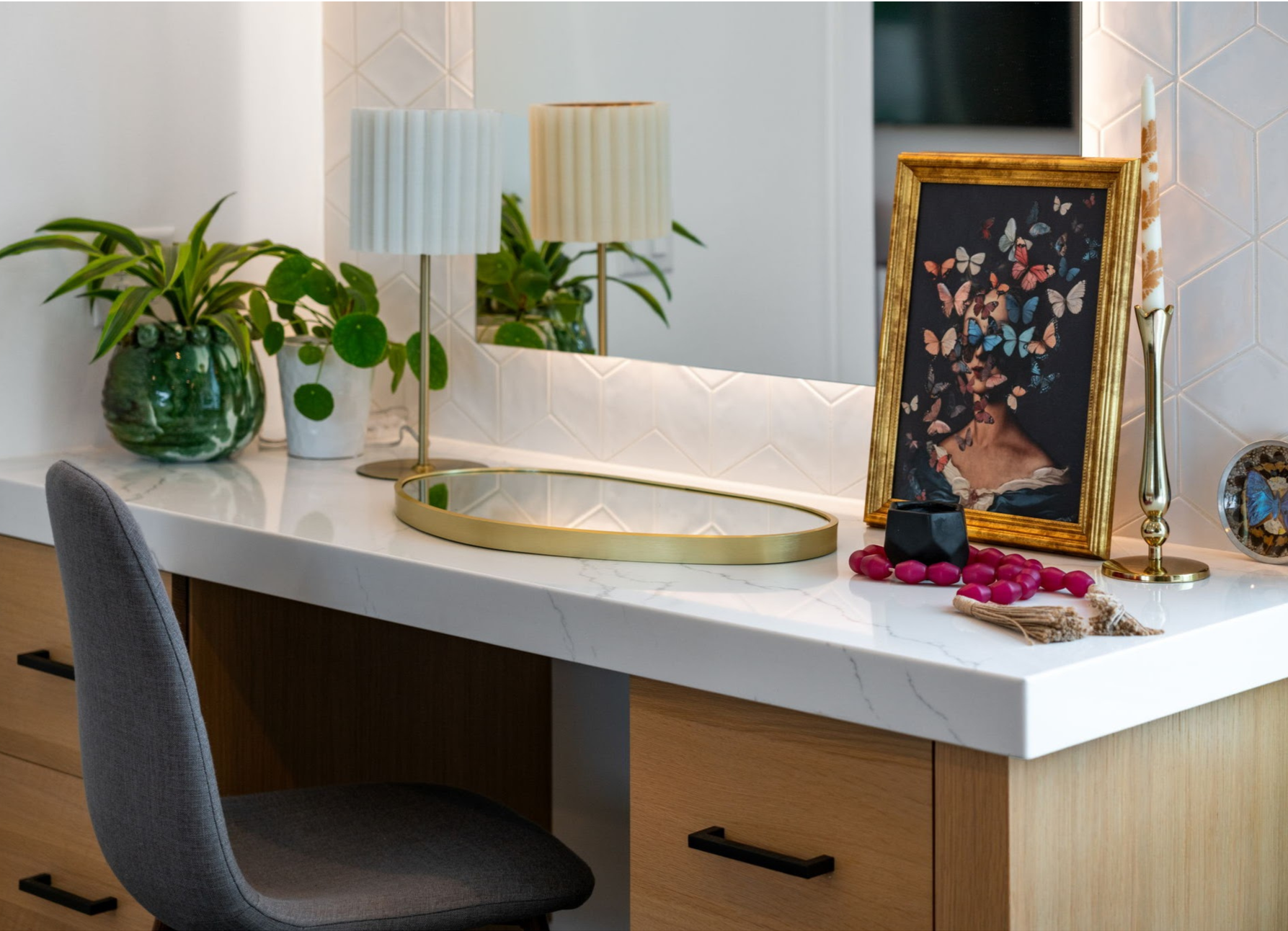 Decorative vanity table with a large mirror, two table lamps, potted plants, a framed butterfly art, a gold tray, and a candlestick, set against a geometric tiled wall.