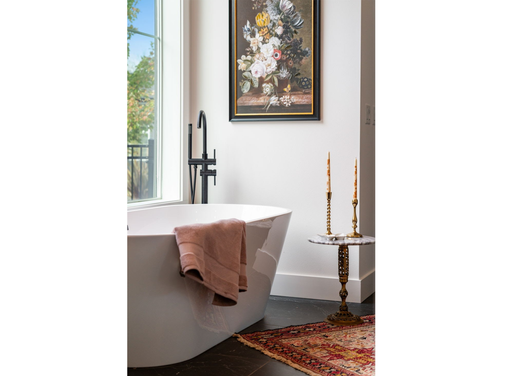 A bathroom with a freestanding white bathtub next to a window, with a pink towel draped over the edge. There is a black floor-mounted bathtub faucet on the left, a framed floral painting on the wall above the tub, and a small round table with two tal
