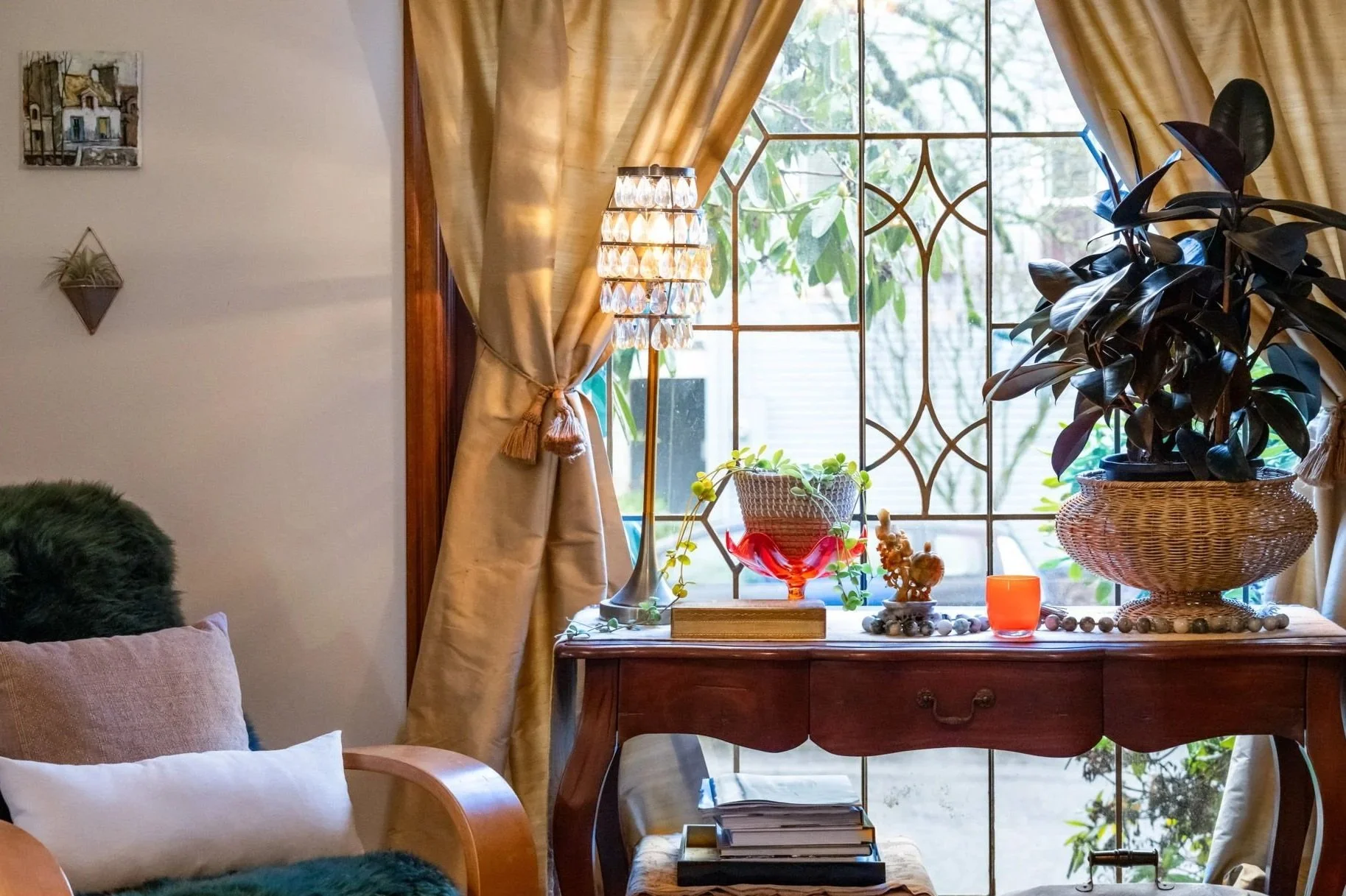 A cozy living room corner with a window, gold curtains, an antique wooden table decorated with plants, candles, and decorative items, a small stool beneath it with books, and a cushioned armchair with pillows and a faux fur covering.