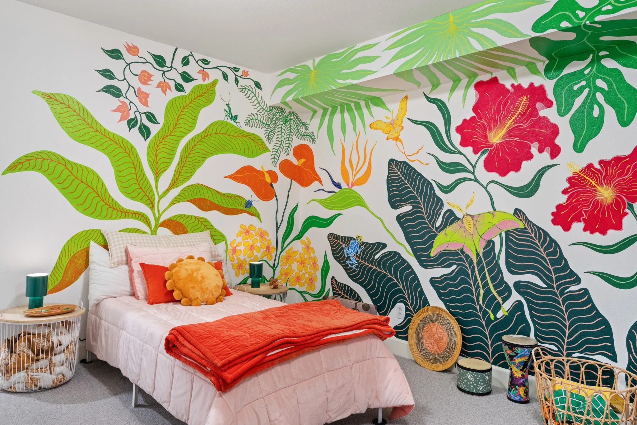 Bedroom with a jungle mural, a bed with a pink blanket and colorful pillows, and various decorative items including lamps, baskets, and musical instruments.