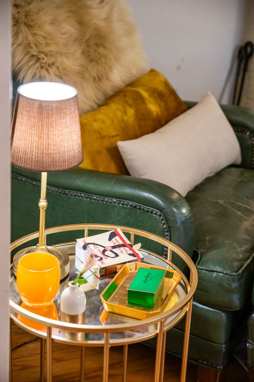 A cozy living room scene featuring a green leather couch with pillows, a side table with a pink lamp, a book, a small vase, and a gold tray with a green box.