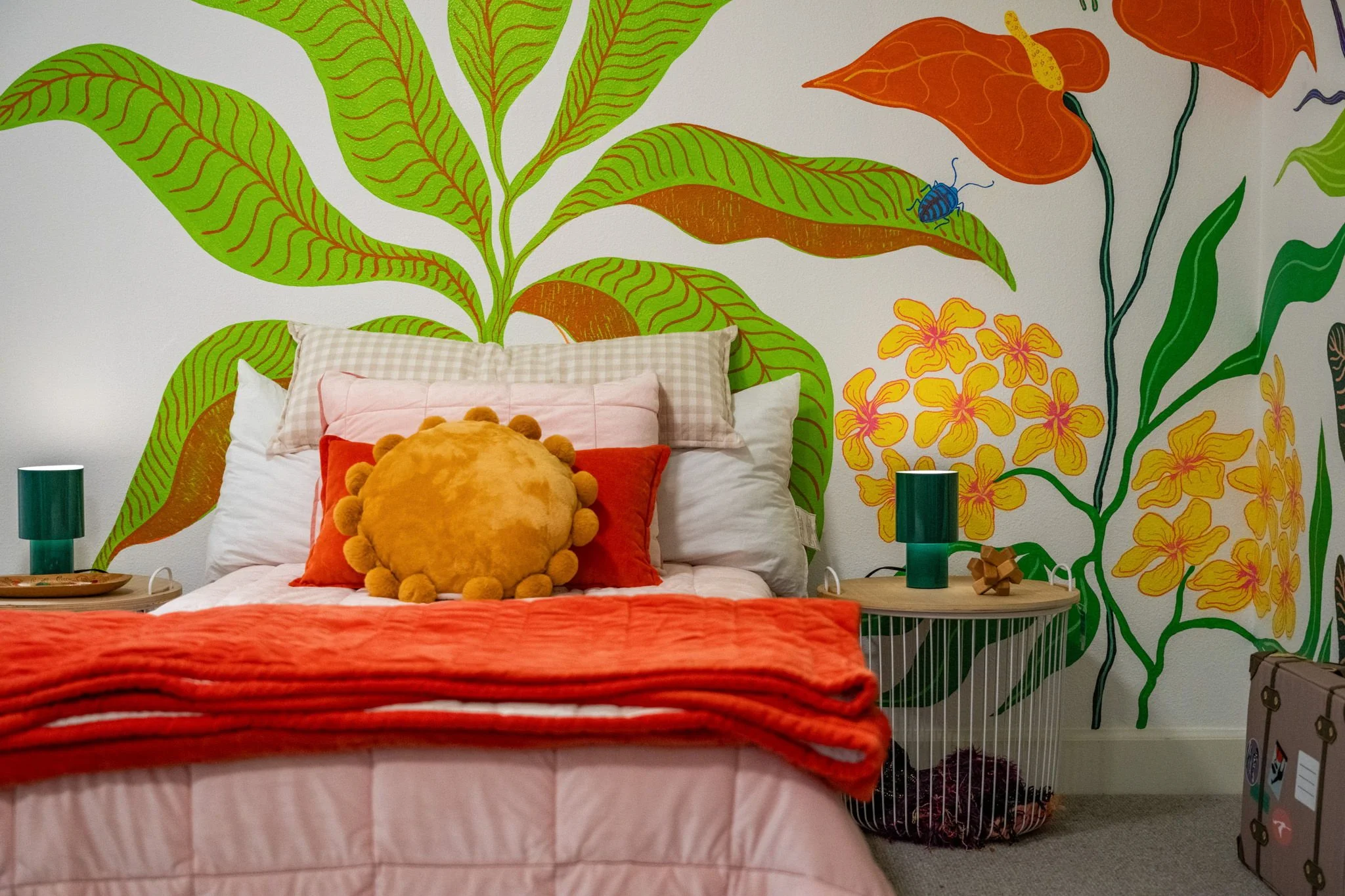 A bedroom with a large, colorful botanical mural on the wall behind the bed. The bed has white and pink linens, with a plush, round yellow pillow with pom-poms. There are green lamps on bedside tables on each side of the bed.