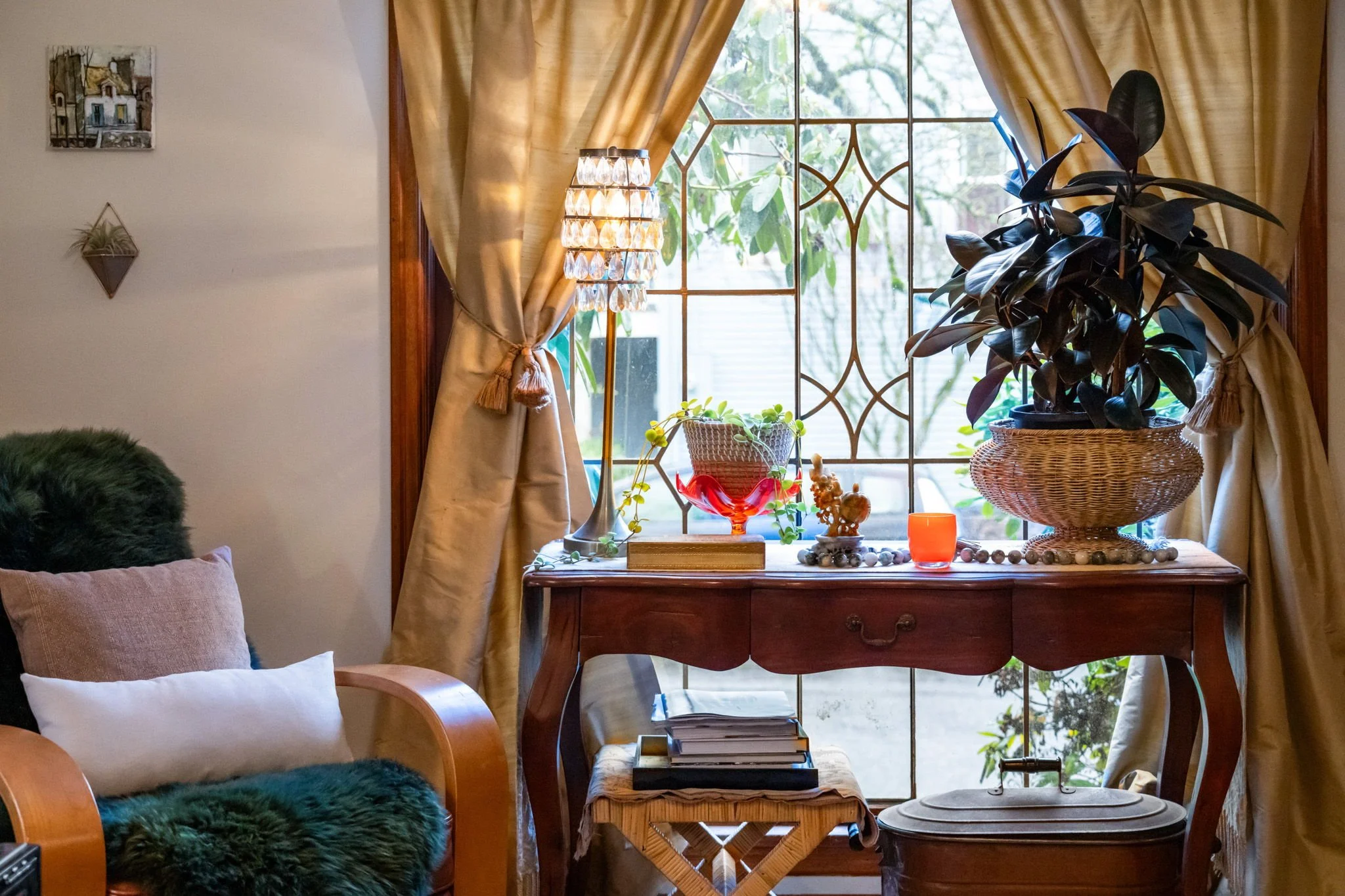 A cozy living room corner with a window, gold curtains, an antique wooden table decorated with plants, candles, and decorative items, a small stool beneath it with books, and a cushioned armchair with pillows and a faux fur covering.