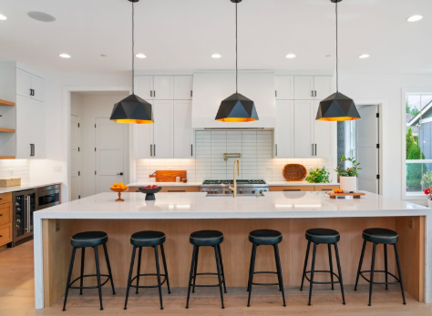 Modern kitchen with white cabinets, a large island with a white countertop, five black bar stools, pendant lights, and a window with a view outside.