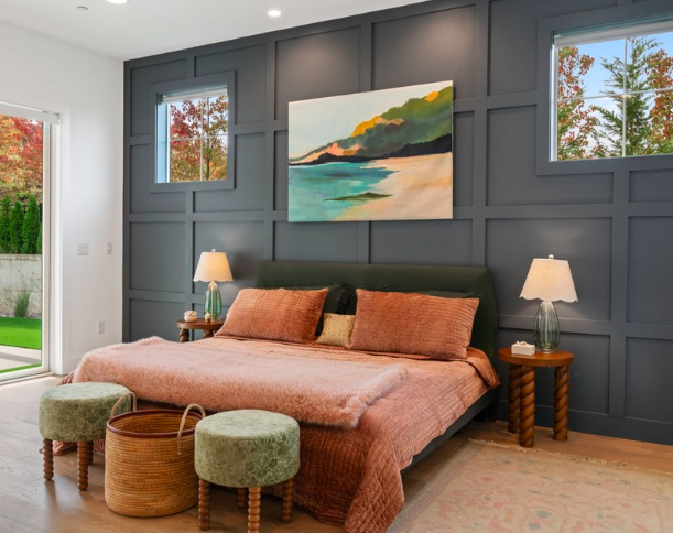 A bedroom with a dark paneled accent wall behind a bed with a green headboard, two table lamps on wooden side tables, and a large abstract landscape painting above the bed. There are two windows on either side of the painting, and a sliding glass doo