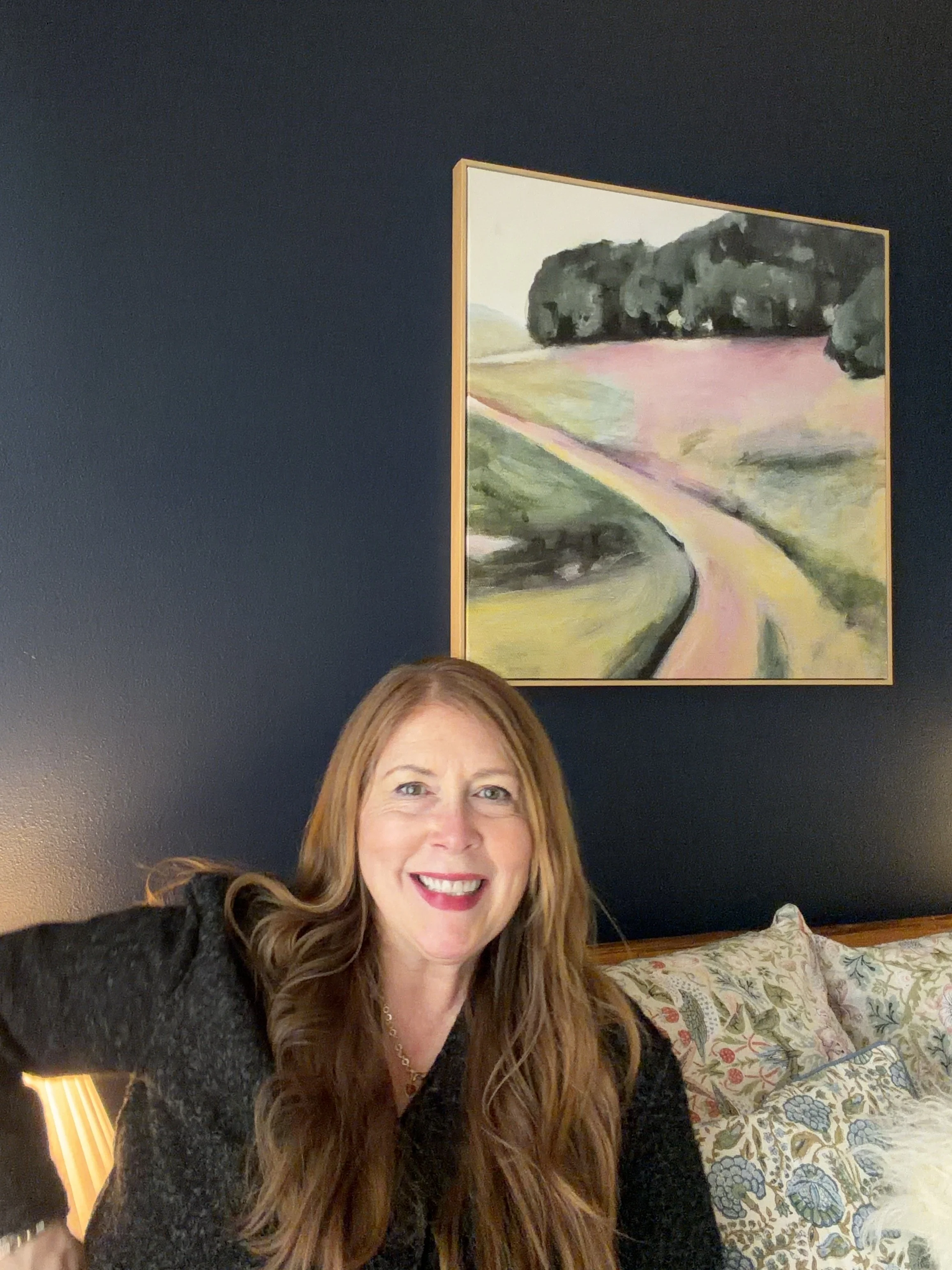 A woman with long red hair smiling, sitting on a bed with patterned pillows, against a dark blue wall with a landscape painting hanging above her.