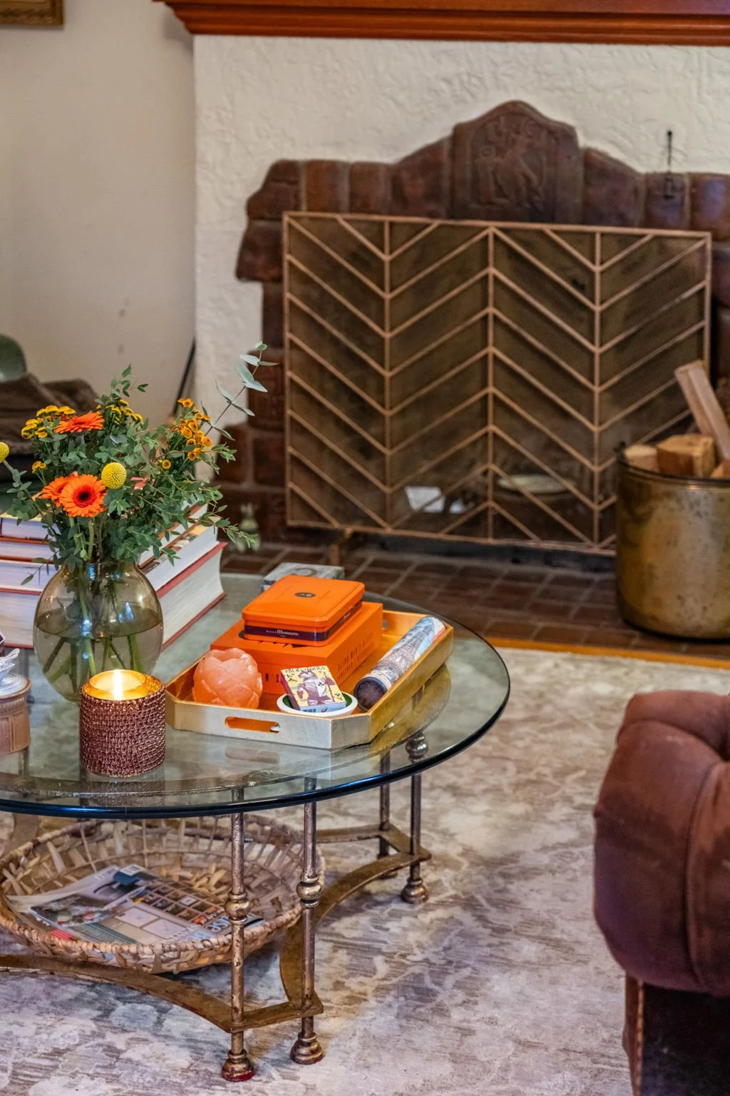 Living room with a glass-top coffee table displaying a vase with flowers, candles, books, and decorative items, in front of a brick fireplace with a wooden screen and a metal bucket filled with firewood.