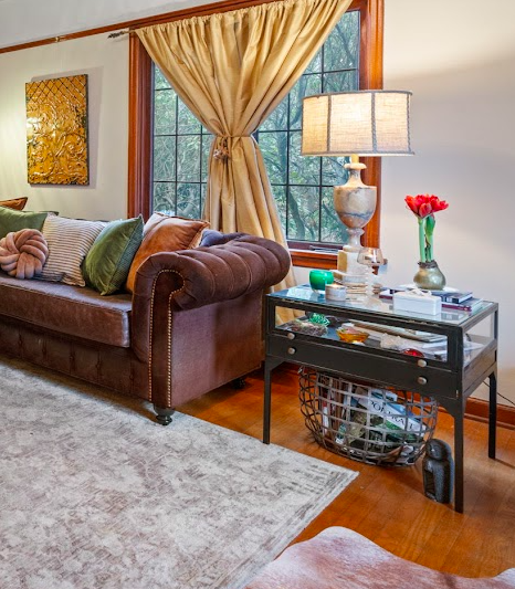 Living room with a brown tufted sofa, gold curtains, a window showing trees outside, a glass side table with a lamp, flower vase, and some books, and a light-colored area rug on hardwood floor.