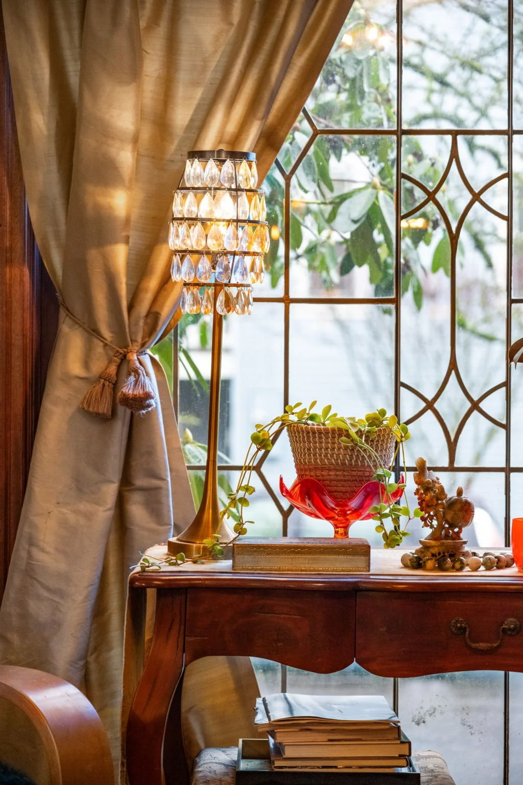 A cozy interior scene featuring a window with decorative ironwork, beige curtains tied back, a table with a red glass bowl holding a potted plant, a small statue, and a brass candlestick, with books stacked underneath.