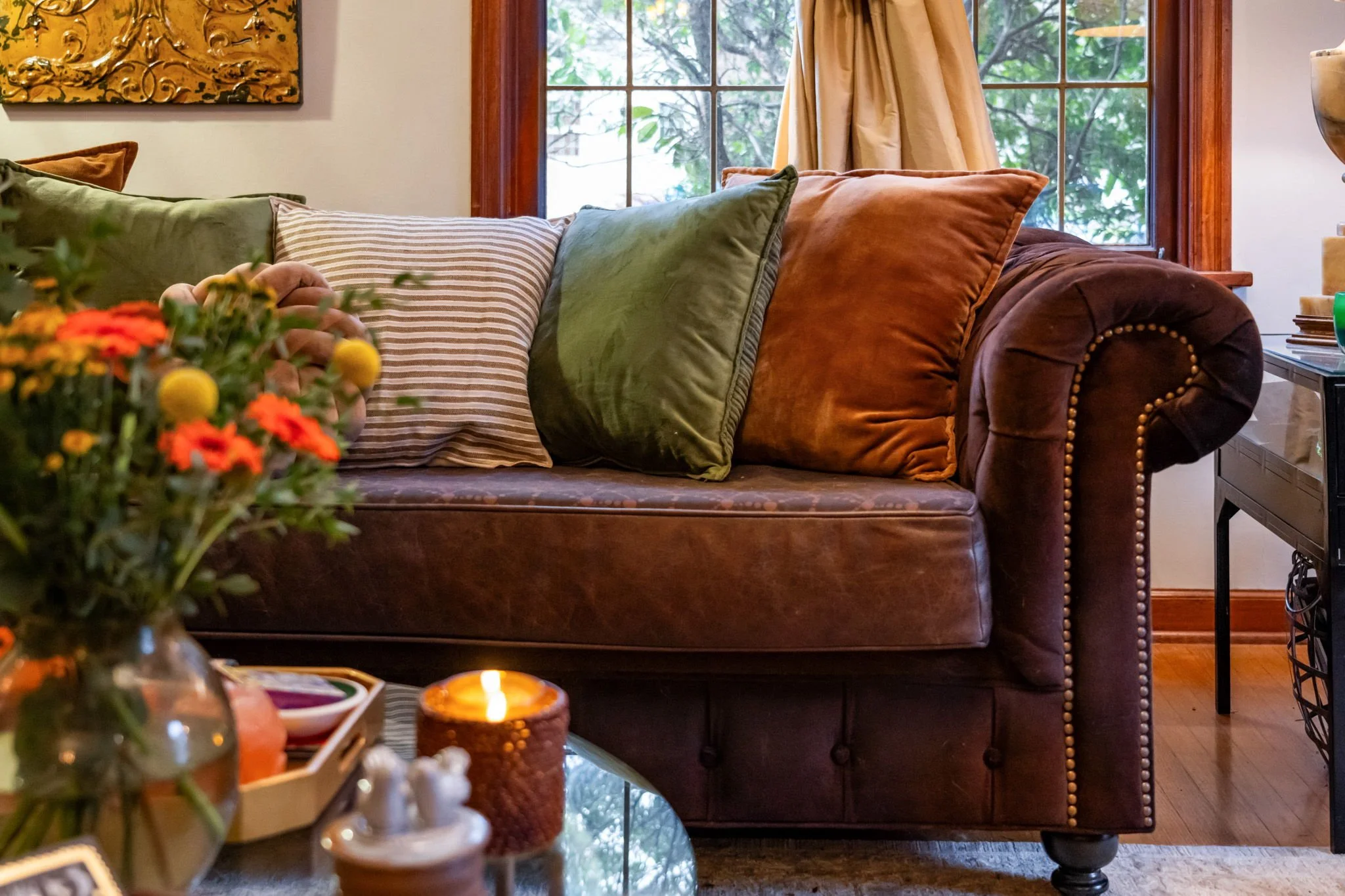 A cozy living room with a brown leather tufted sofa holding several pillows in shades of green, orange, and a striped pattern. In front, a glass coffee table displays flowers in a vase, a lit candle, and other small decorative items. A window with be