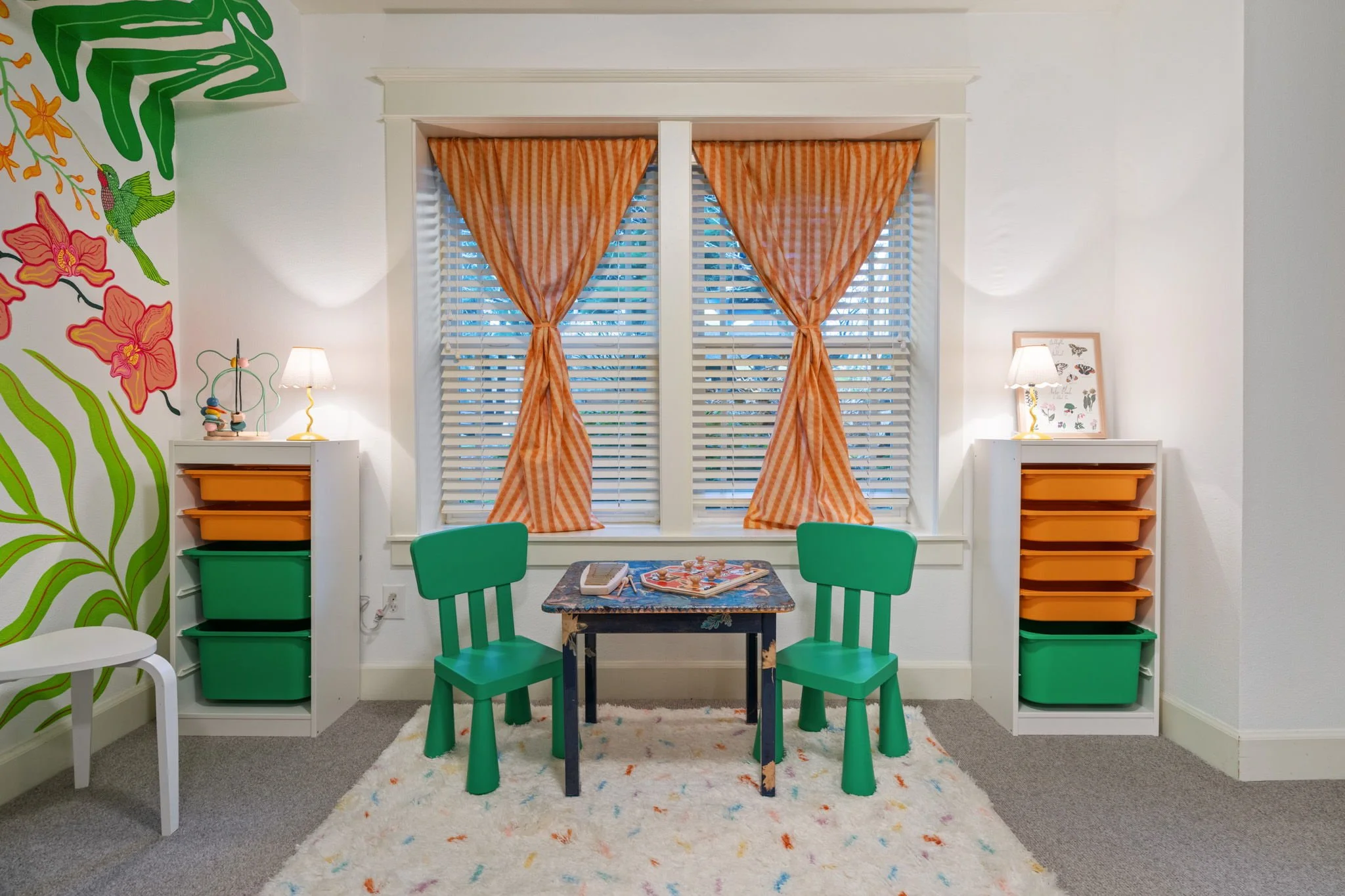 A children's playroom with a window featuring orange checked curtains, a small table, two green chairs, matching shelving units with orange and green storage bins, a white bench, colorful wall mural with floral and botanical designs, and a patterned 