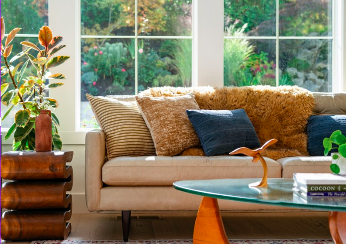 Living room with beige sofa, multiple textured and striped pillows, a glass coffee table, a wooden side table with a potted plant, large windows showing a garden with trees and bushes.