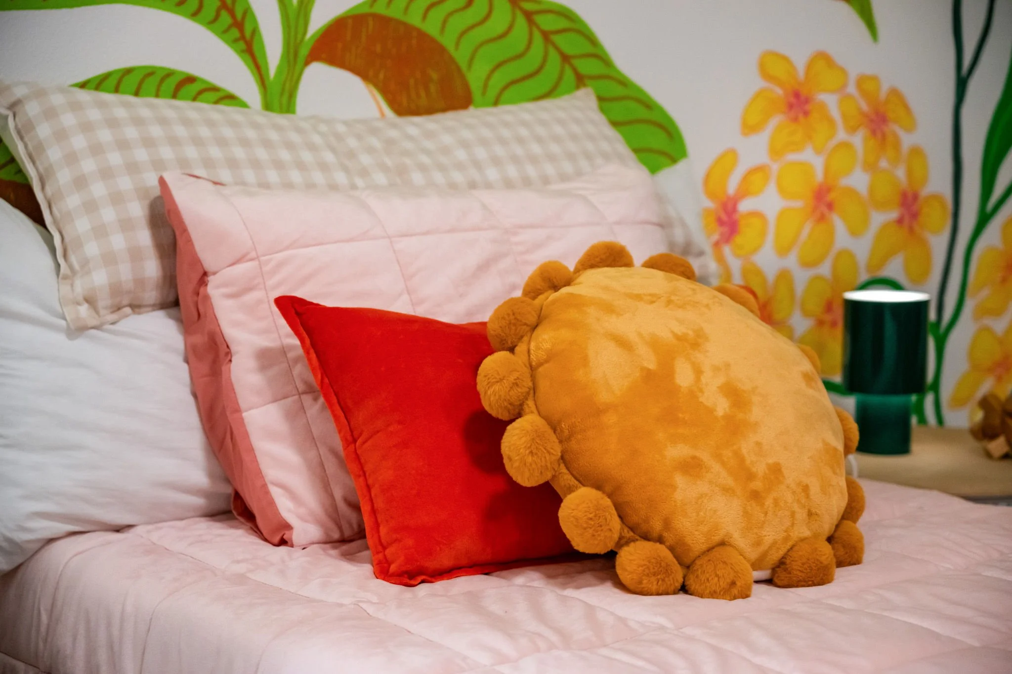 Decorative bed with pillows, including a fluffy yellow round pillow with pom-pom trim, against a vibrant mural with green leaves and yellow flowers.