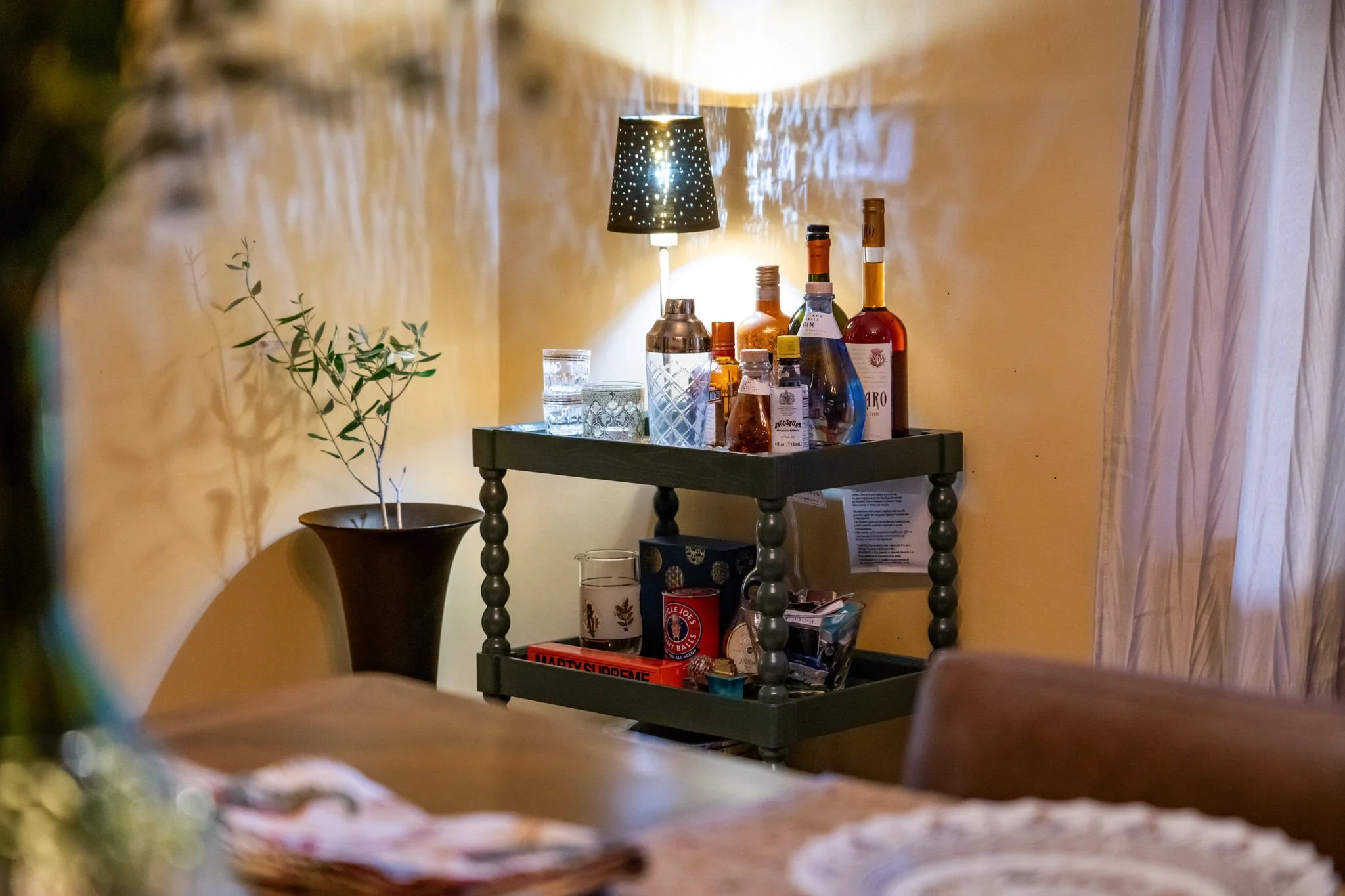 A bar cart with bottles of alcohol, glasses, and a lamp in a cozy room.