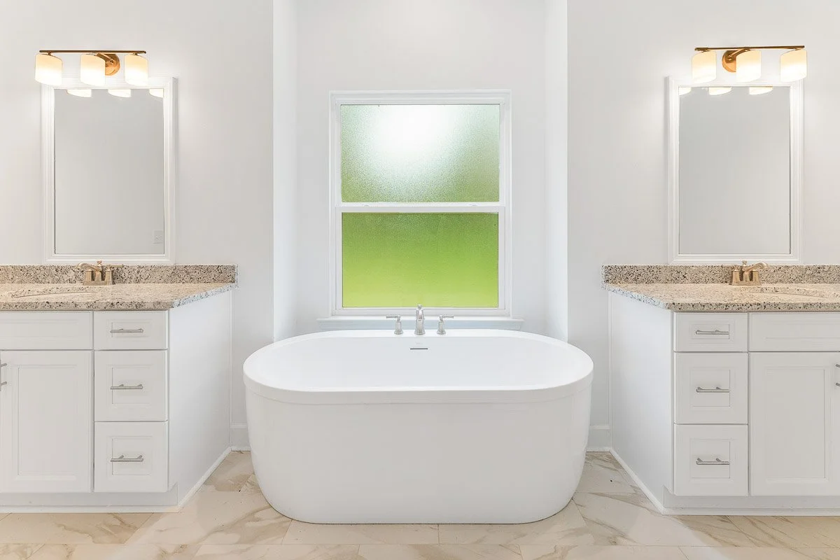 Real Estate photo of a bright bathroom with dual vanities, granite countertops, mirrors, modern lighting, a freestanding bathtub, and a frosted window.