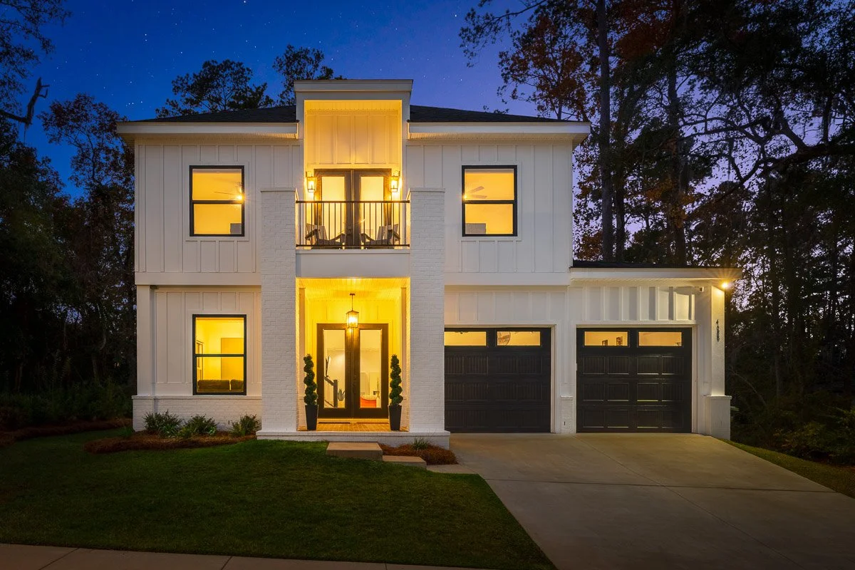 Real Estate photo of a modern two-story house with white exterior and black garage doors illuminated at night, surrounded by trees and a well-kept lawn.