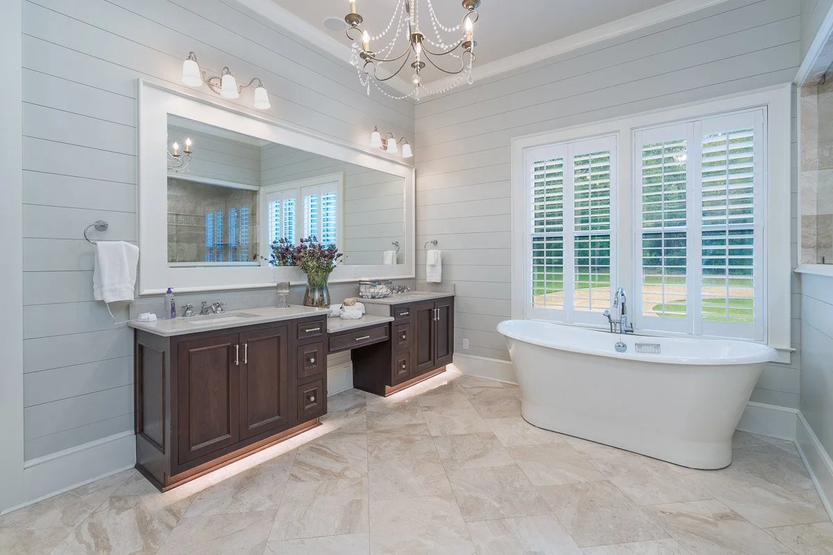 Real Estate photo of a modern bathroom with a white bathtub near a large window with plantation shutters, a dark wood vanity with a large mirror, a chandelier, and neutral floor tiles.