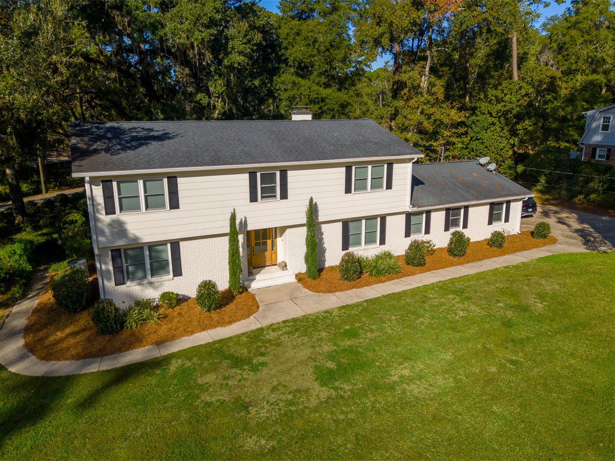Aerial Real Estate photo of a mid-century, two-story home with white exterior walls, black shutters, and a dark gray roof, surrounded by a well-maintained lawn, trees, and shrubs.