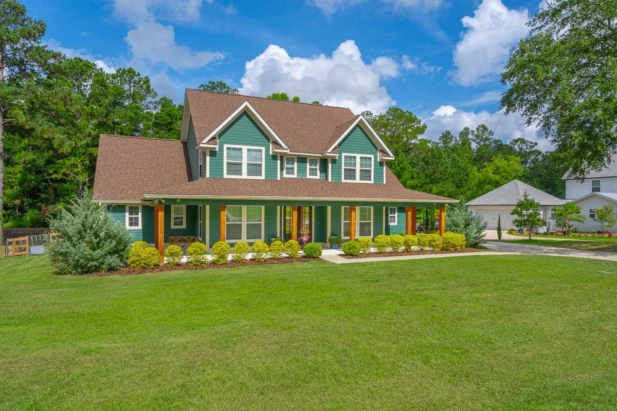 Real Estate photo of a large, modern two-story house with a front porch, surrounded by a well-maintained green lawn and landscaped bushes, under a partly cloudy blue sky.