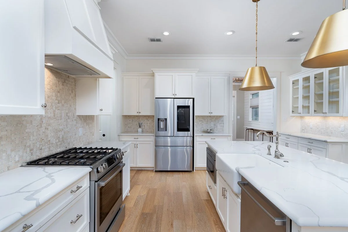 Bright white kitchen with stainless steel refrigerator, oven, and modern cabinetry, with gold pendant lights overhead and hardwood floors.