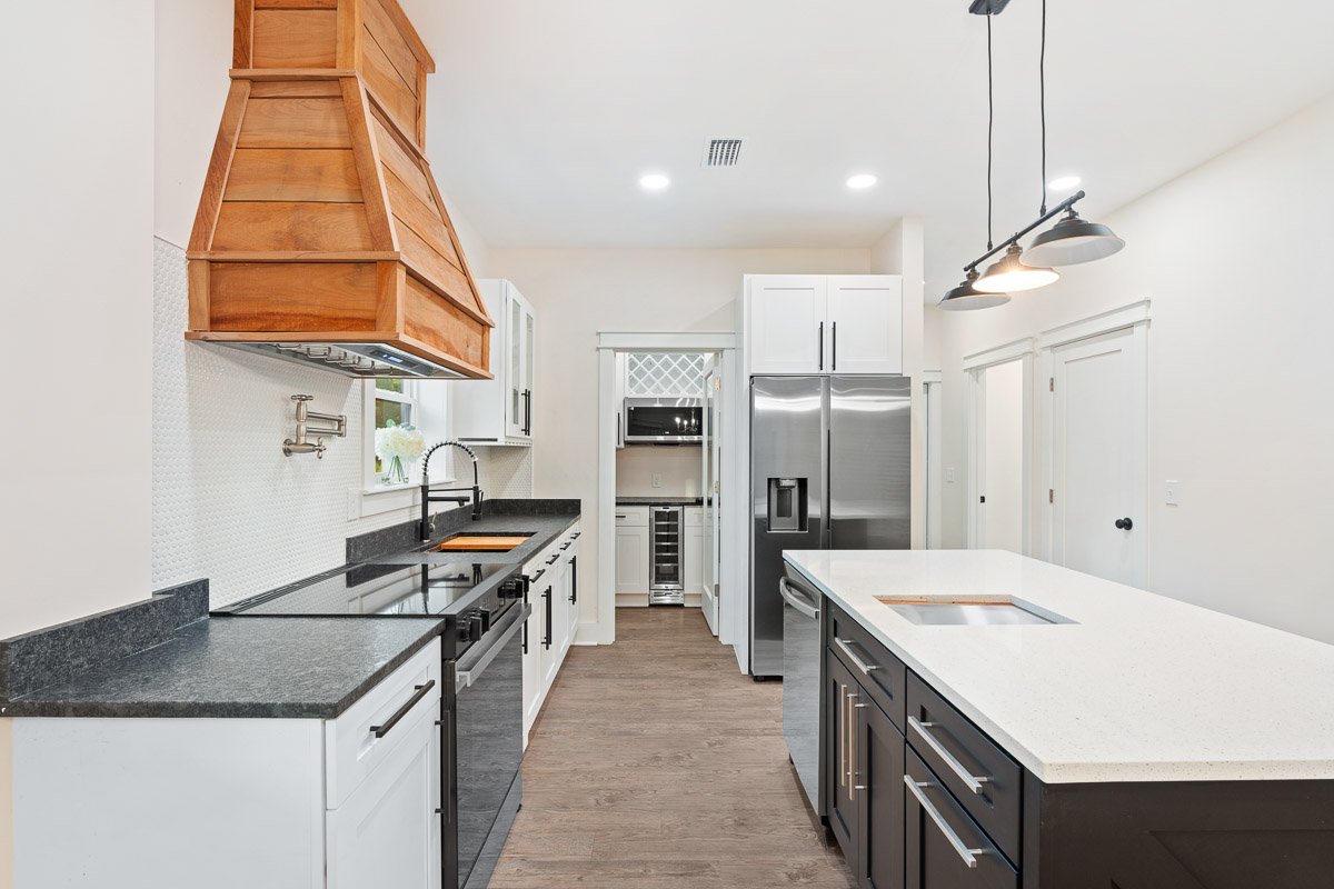 Real Estate photo of a modern kitchen with white walls, black and white cabinets, a white island, stainless steel appliances, and a wooden range hood.