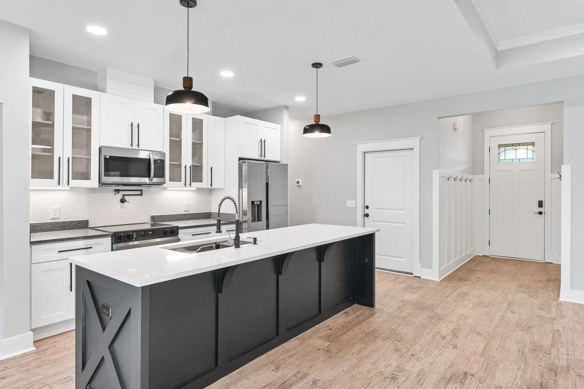 Real Estate photo of a newly renovate modern kitchen with white cabinets, a stainless steel refrigerator, microwave, and stove, a black kitchen island with a white countertop, hardwood floors, pendant lighting, and a small entryway with hooks.