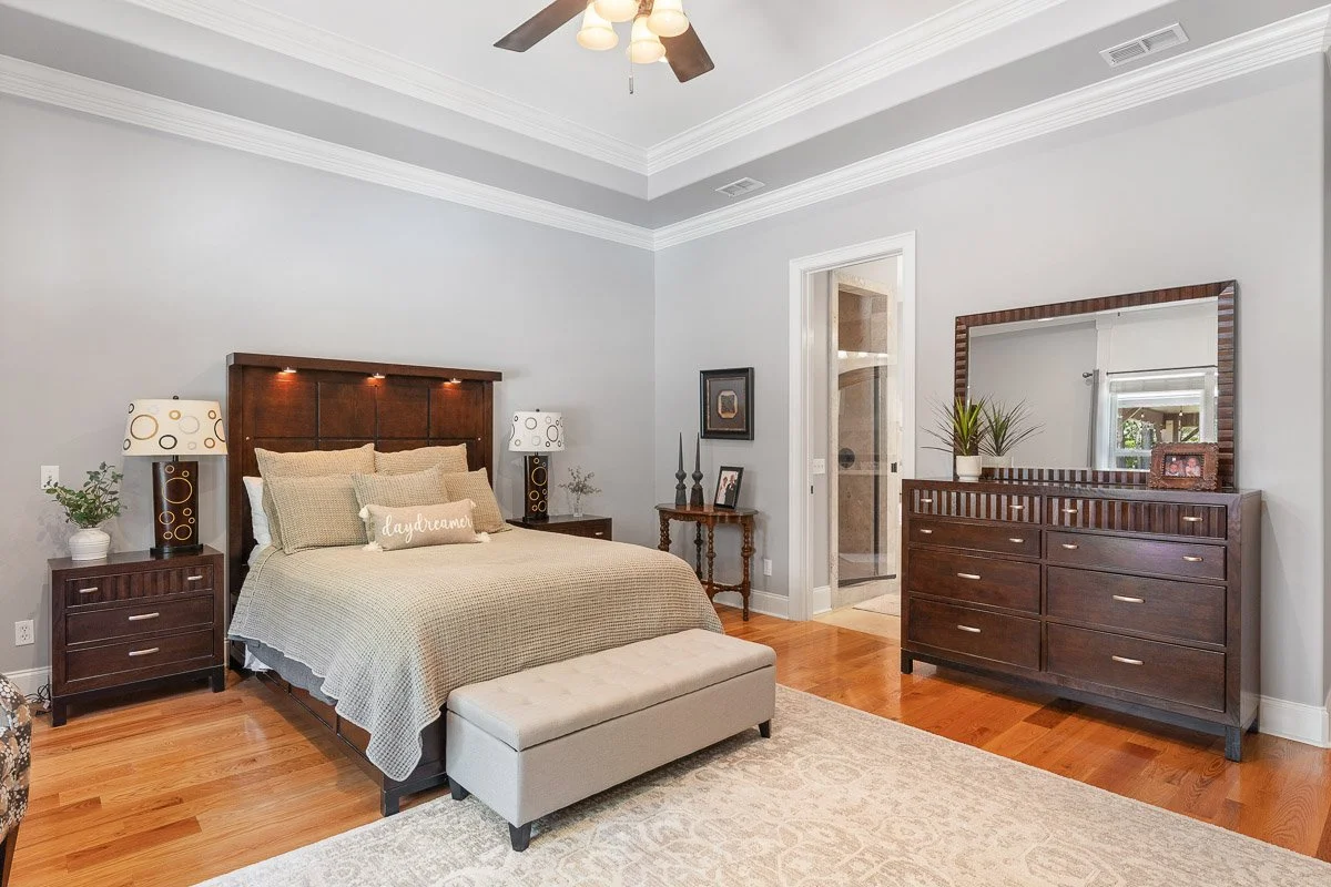Real Estate photo of a bedroom with a large bed, nightstands with lamps, a dresser with mirror, a bench at the foot of the bed, and a doorway leading to a bathroom with a shower.