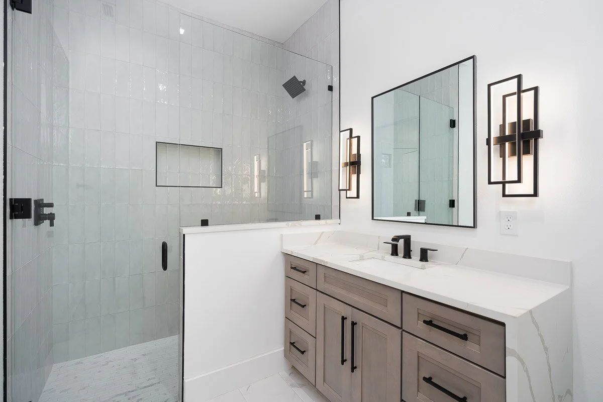 Modern bathroom with white tiled shower area, glass door, grey vanity with black hardware, large mirror, and contemporary black wall sconces.