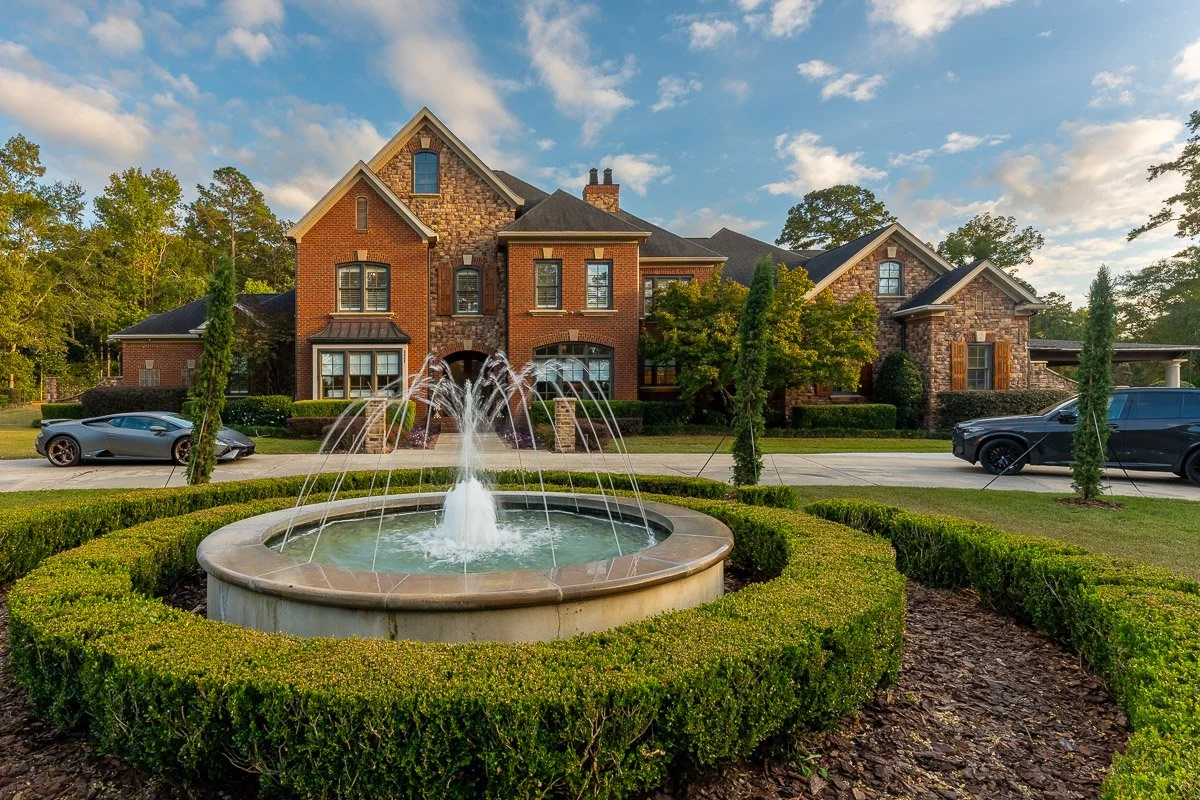 Real Estate photo of a large brick house with multiple gables, surrounded by trees and a well-manicured lawn. A fountain with water jets is in the front yard, and two black cars are parked on the driveway.