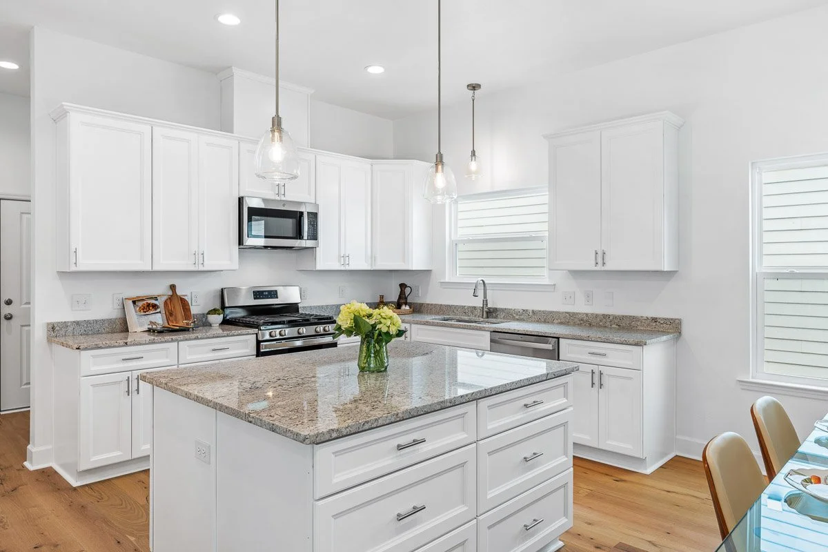 Real Estate photo of a modern white kitchen with granite countertops, island, stainless steel appliances, pendant lighting, and a dining table with chairs.