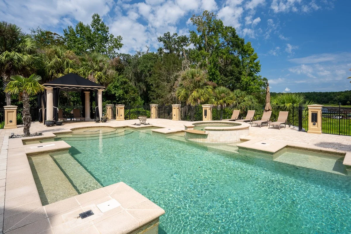 Real Estate photo of a luxurious outdoor swimming pool with a hot tub, surrounded by lounge chairs, a gazebo, green trees, and a clear blue sky.