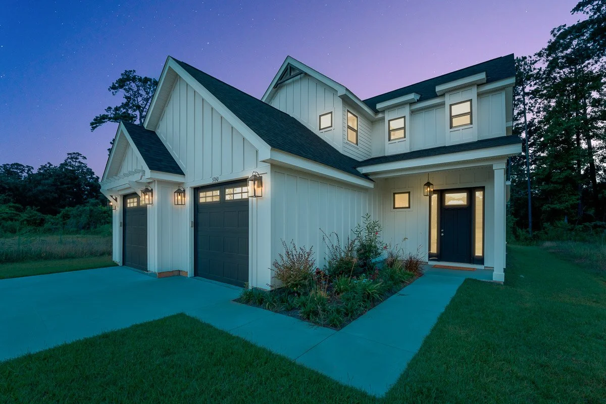 Real Estate photo of a modern two-story white house with black garage doors and black front door, illuminated by exterior lantern lights during dusk, surrounded by a green lawn and trees in the background.
