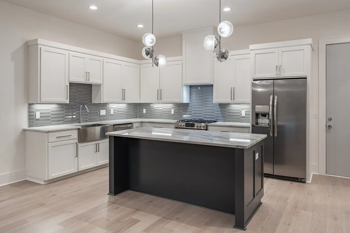 Real Estate photo of a modern kitchen with white cabinets, long gray tile backsplash, stainless steel appliances, and a black island with a marble countertop.