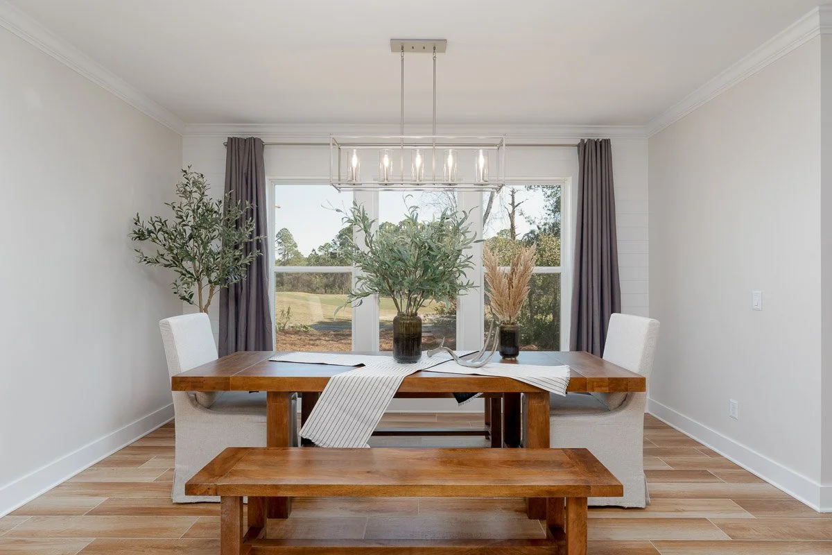 A dining room with a wooden table, white chairs, a bench, and a large window with gray curtains. The table has vases with greenery and pampas grass, and a modern chandelier hangs above.