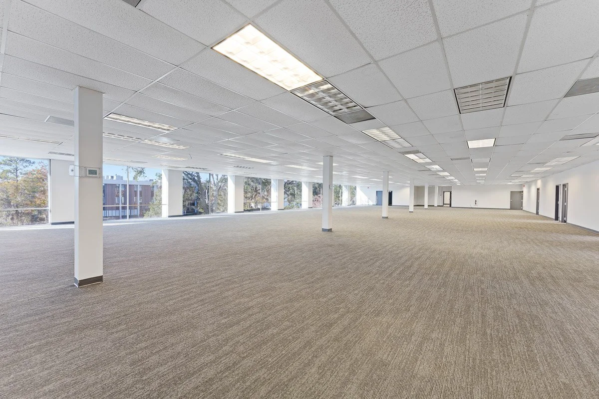 Commercial Real Estate photo of a large empty office space with carpeted floor, white walls, and large windows letting in natural light.