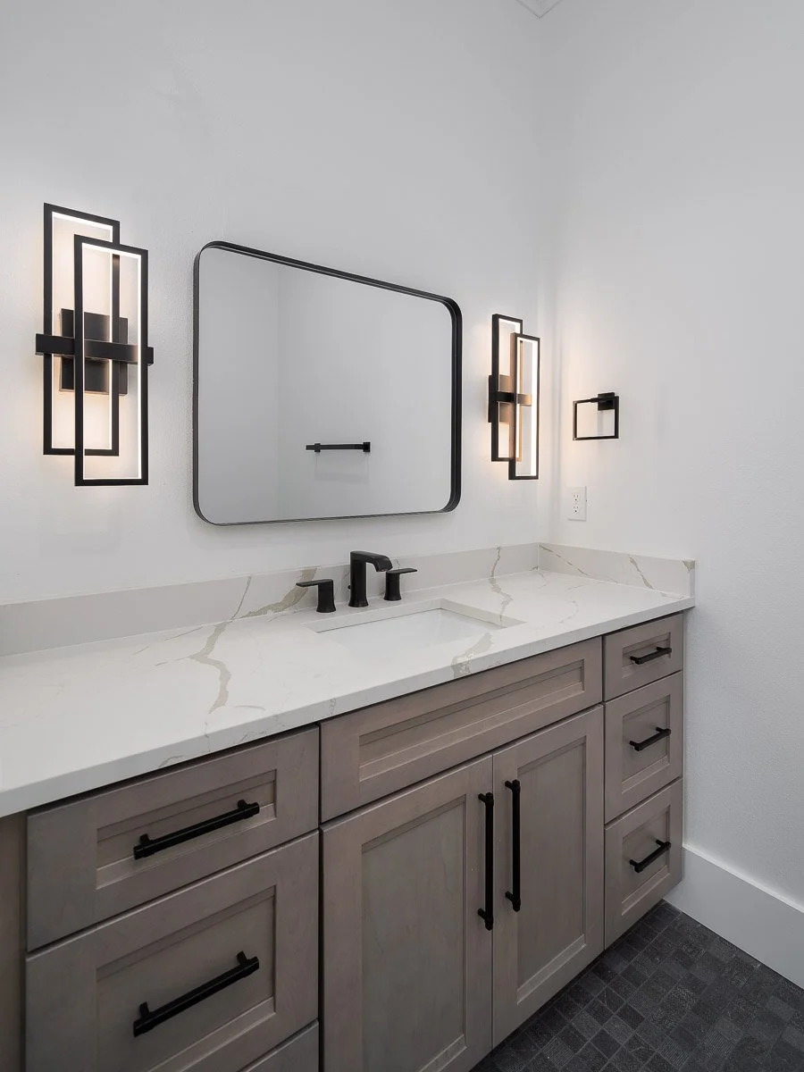 A modern bathroom vanity with a white marble countertop, gray cabinetry with black handles, a square mirror, black wall sconces with frosted glass, and a black faucet.