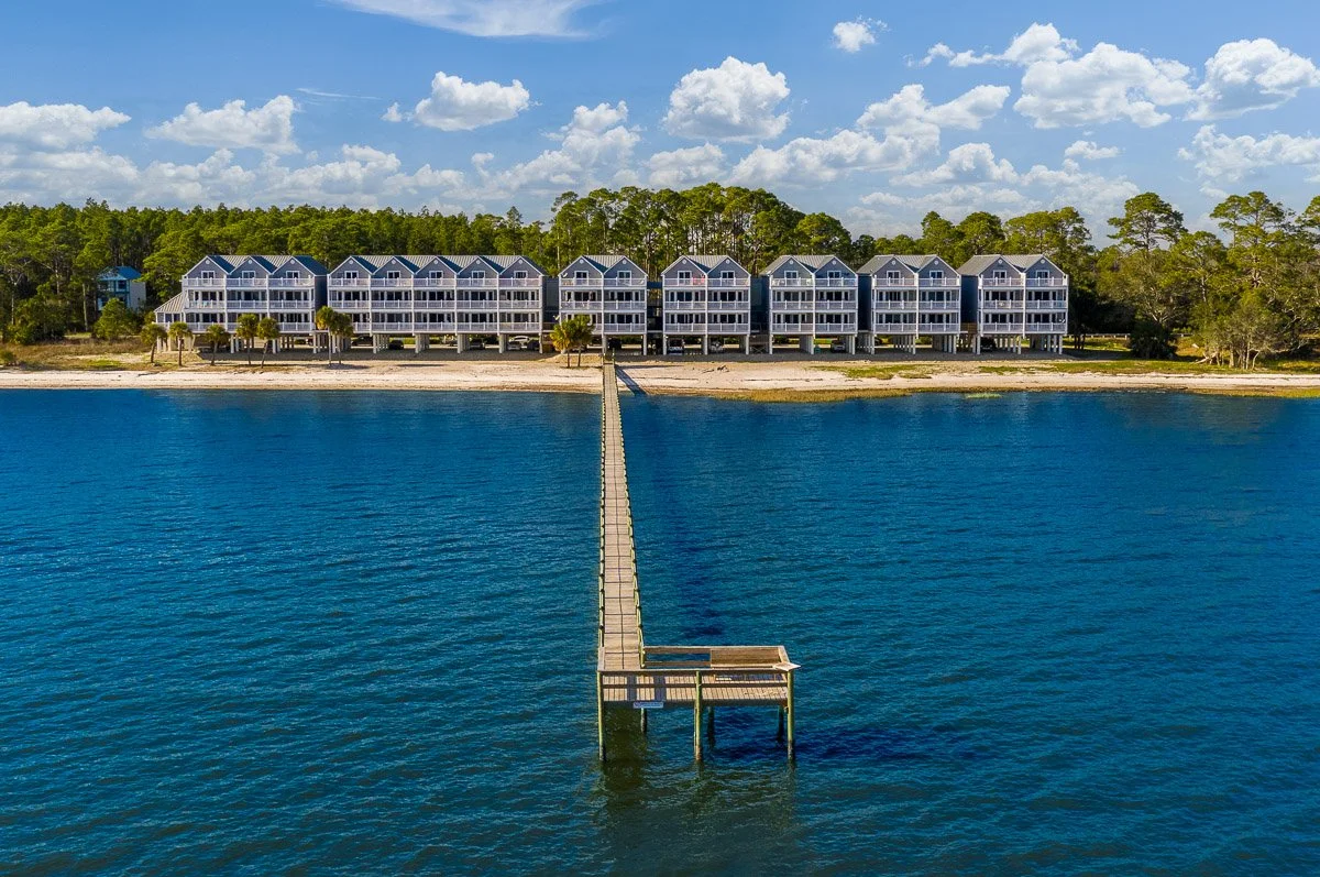 Aerial real Estate photo of a row of beach condos elevated on stilts with a long pier extending into the water in front of them, and a partly cloudy sky in the background.