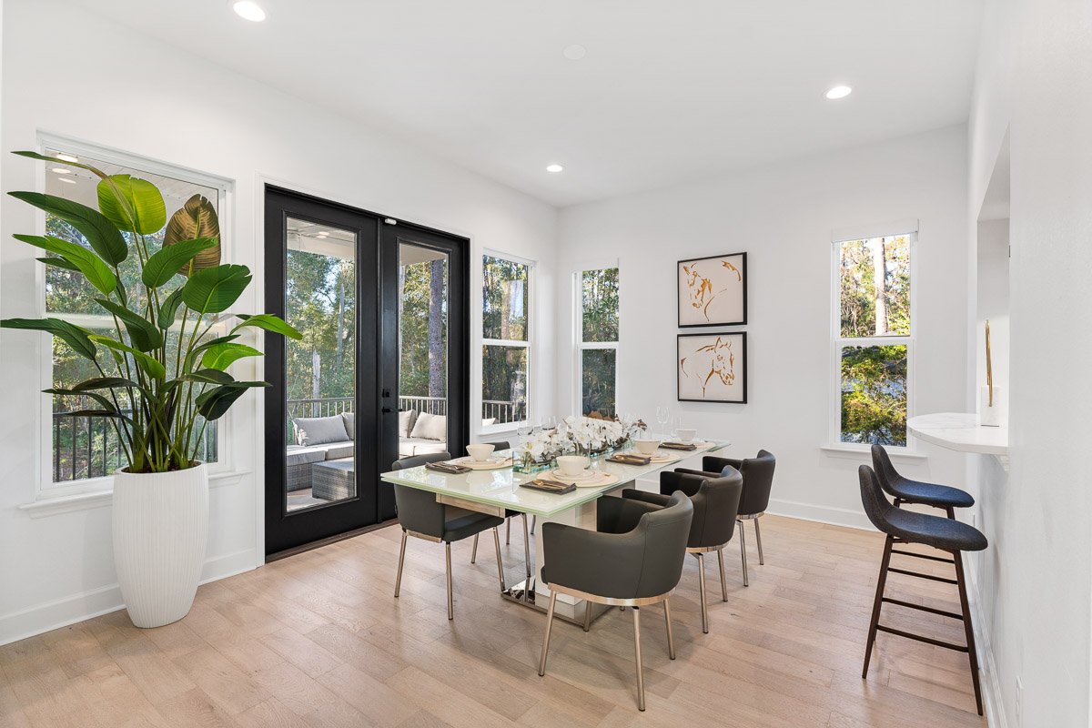Real Estate photo of a bright dining room with large black-framed glass doors, white walls, a rectangular dining table with six dark chairs, and a large potted plant near the window.