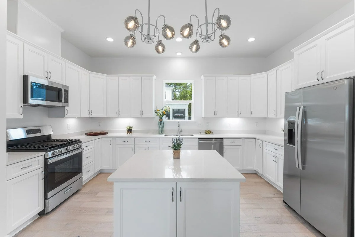 Real Estate photo of a bright white kitchen with a central island, stainless steel appliances, and white cabinets. A window above the sink shows a view outside. There is a modern chandelier hanging from the ceiling.