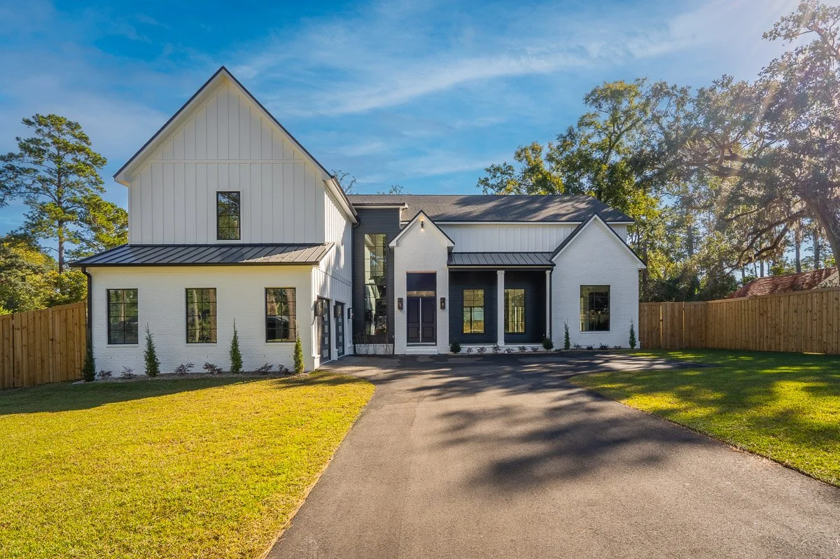 Modern white house with dark accents, large front yard, and wooden fence, under a blue sky with some clouds and trees in the background.