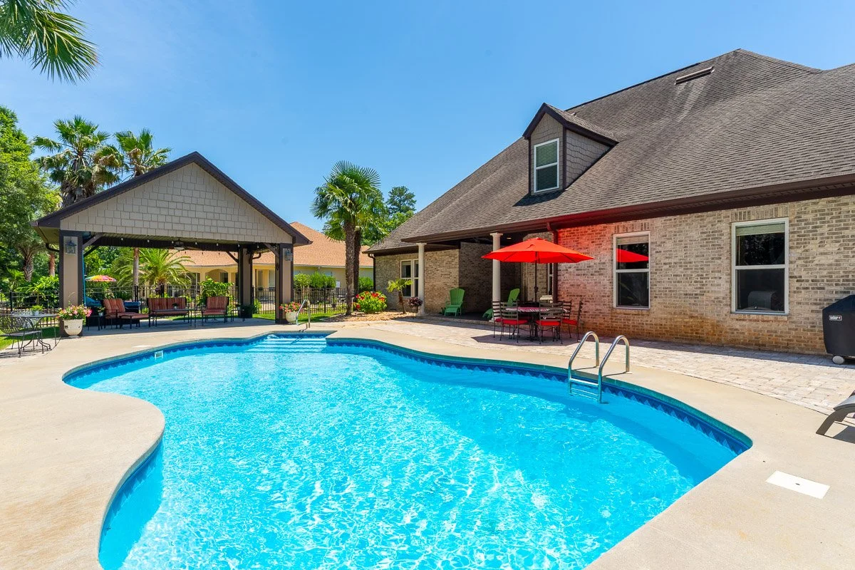 Real Estate photo of a backyard with a swimming pool, patio furniture, and a covered pavilion, surrounded by trees and a bright blue sky.