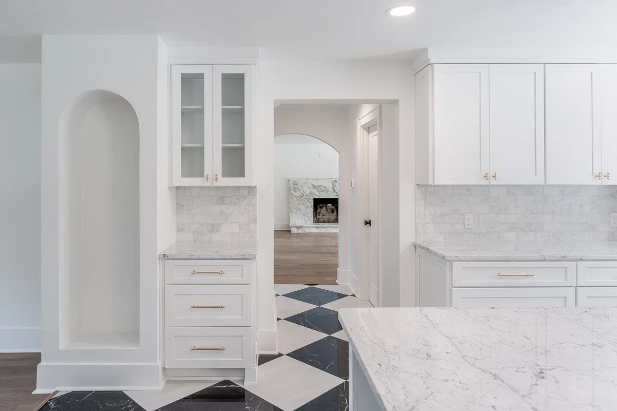 Architectural style Real Estate photo of a modern white kitchen with marble countertops, gold hardware, glass-front cabinets, and diagonally patterned black and white tile floor, view into living room with fireplace.