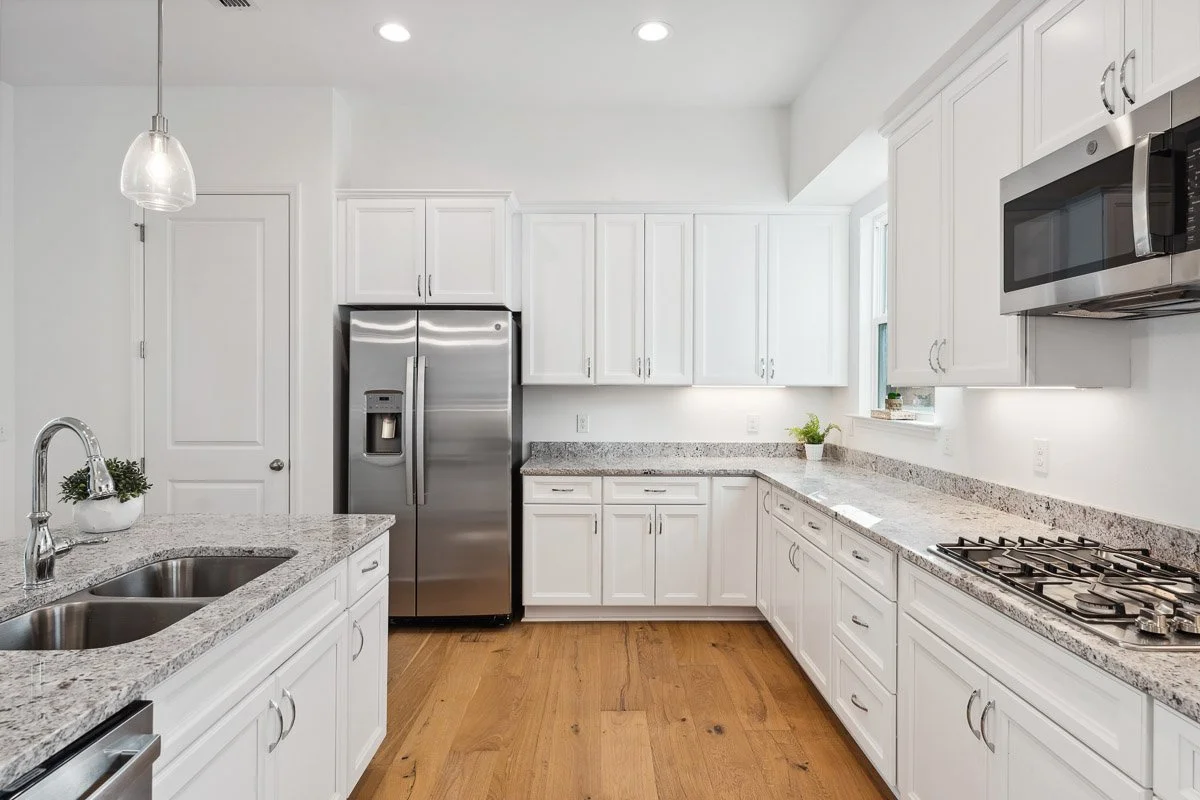 Real Estate photo of a clean white kitchen with granite countertops, stainless steel appliances, a double sink, and a hardwood floor. There is a window above the counter, and a pendant light hanging from the ceiling.
