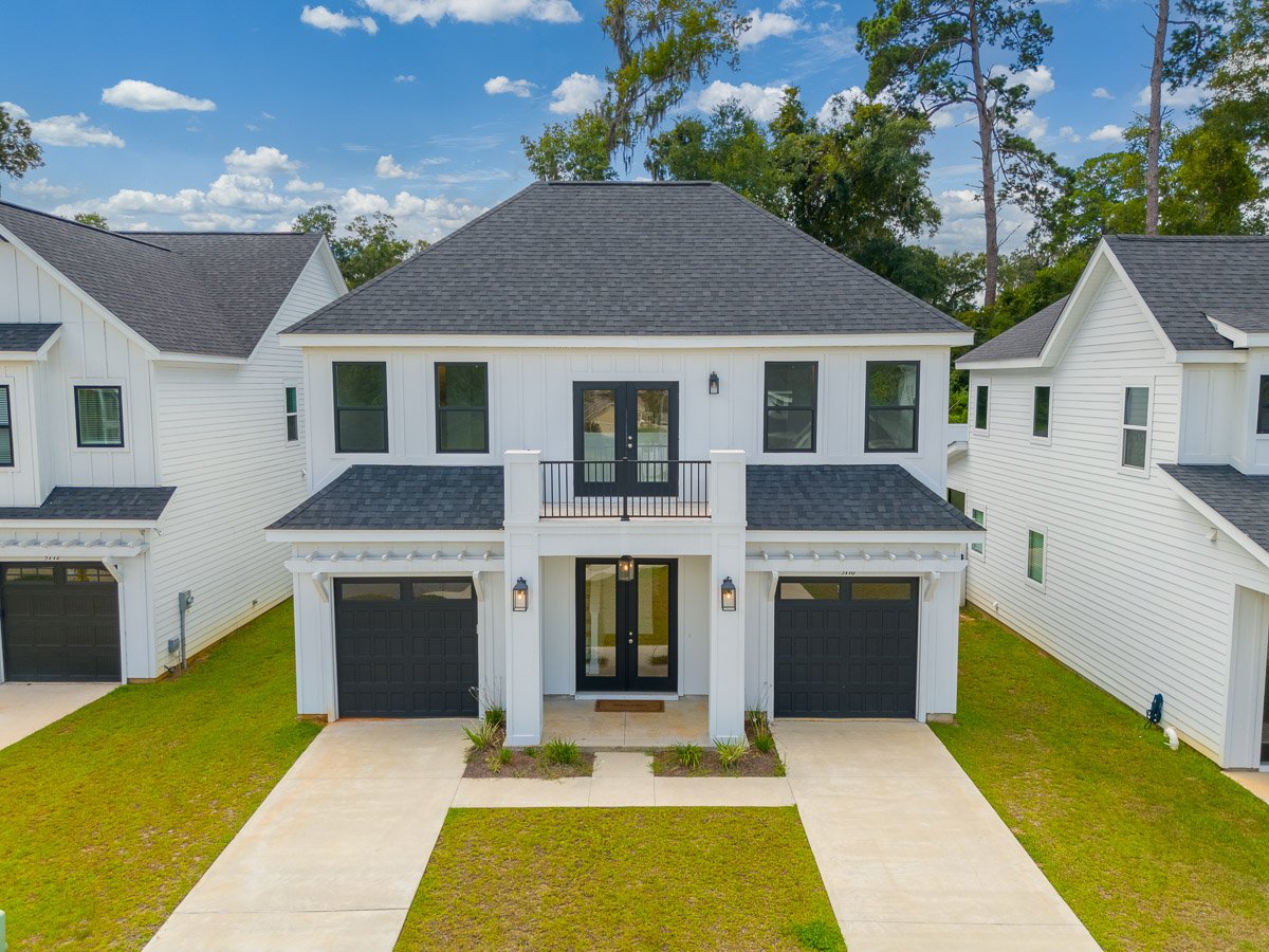 Low angle aerial Real Estate photo of a modern two-story house with white exterior and black garage doors, situated between two similar houses, with a concrete driveway, small front yard, and a balcony above the front door, under a partly cloudy sky.