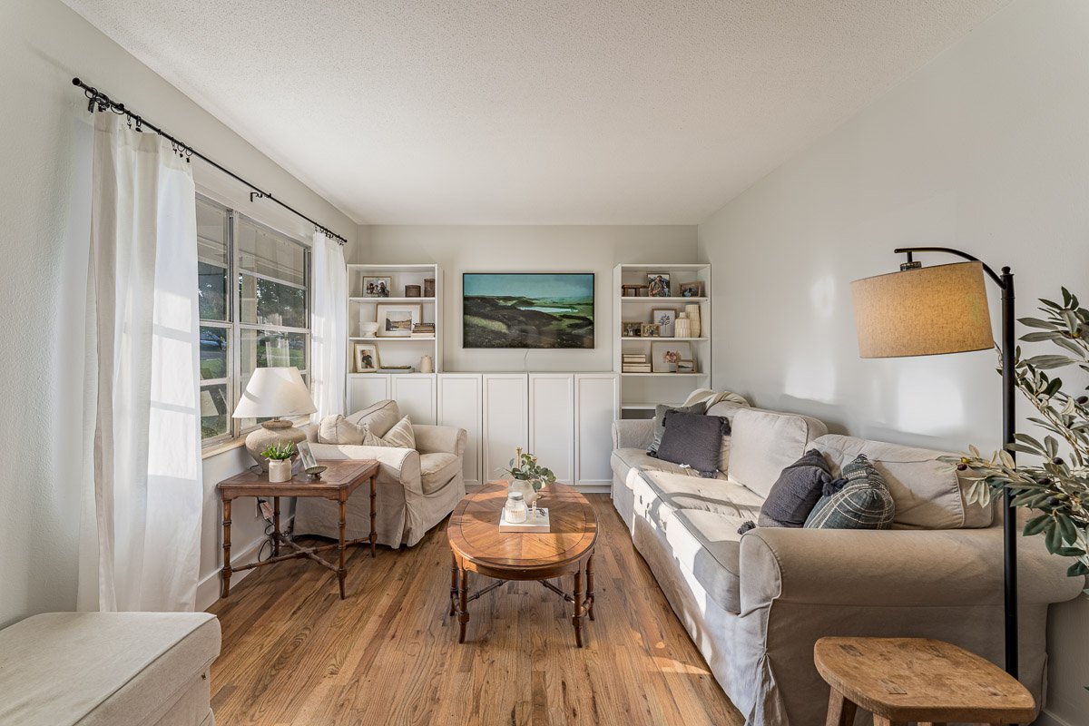 Real Estate photo of a cozy living room with a large window, white curtains, and hardwood floors. It features a beige sofa with black and plaid pillows, a wooden coffee table, a side table with a lamp, a tall floor lamp, and built-in white shelves wi