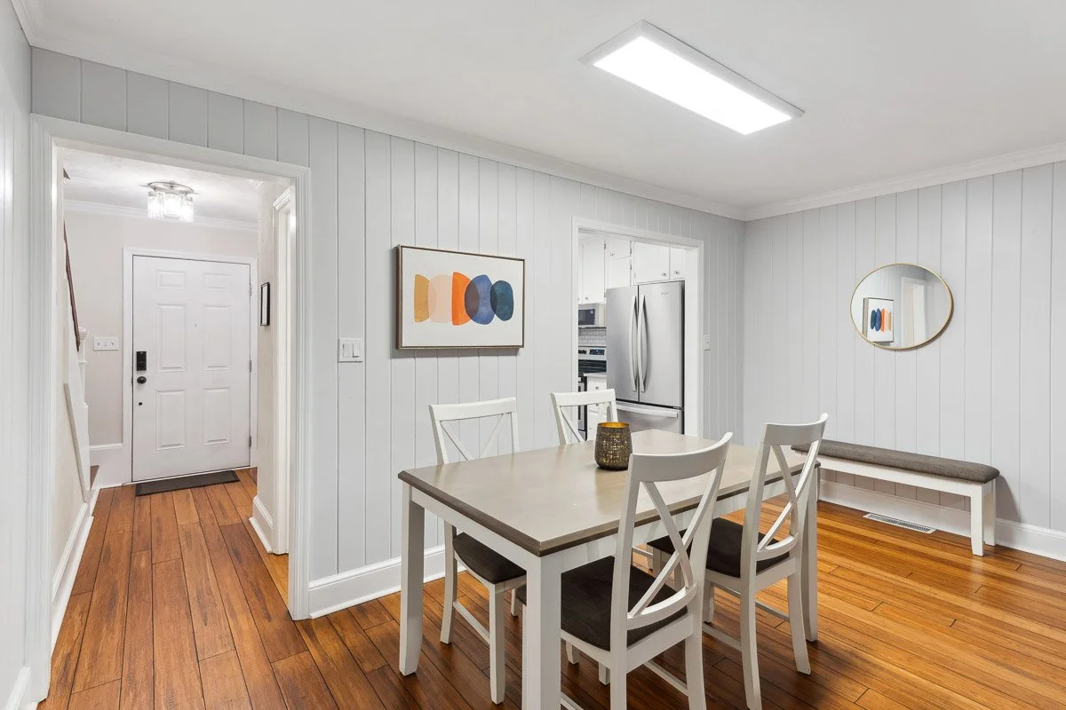 Real Estate photo of a dining room with white paneled walls, hardwood floor, a rectangular table with four chairs, a bench with gray cushion, and wall art with colorful abstract shapes.